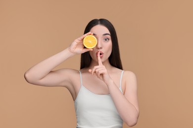 Beautiful young woman with piece of orange showing hush gesture on beige background