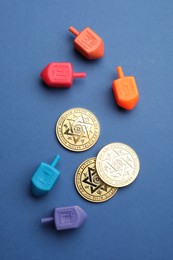 Photo of Dreidels with Jewish letters and coins on blue background, flat lay. Traditional Hanukkah game