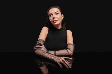 Photo of Beautiful young woman in evening gloves at black glass table against dark background
