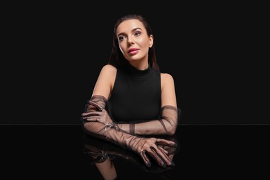 Beautiful young woman in evening gloves at black glass table against dark background