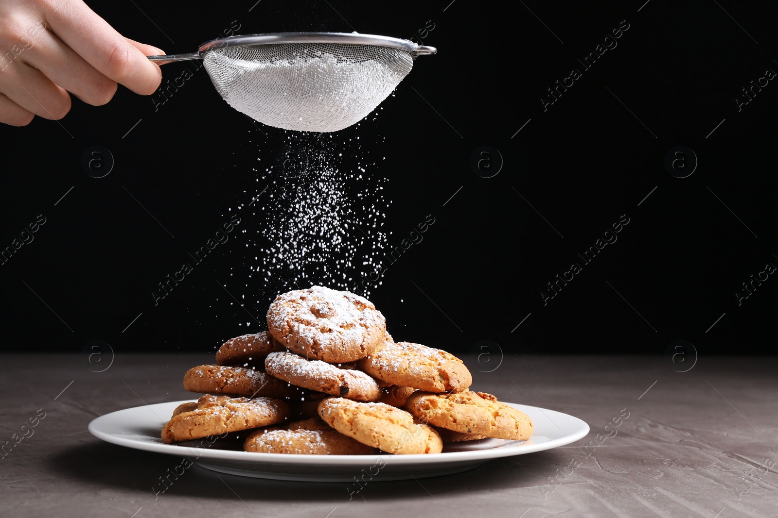 Photo of Woman with sieve sprinkling powdered sugar onto cookies at grey textured table, closeup