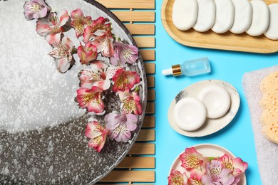 Photo of Bowl of water with flowers and different spa supplies on light blue background, flat lay