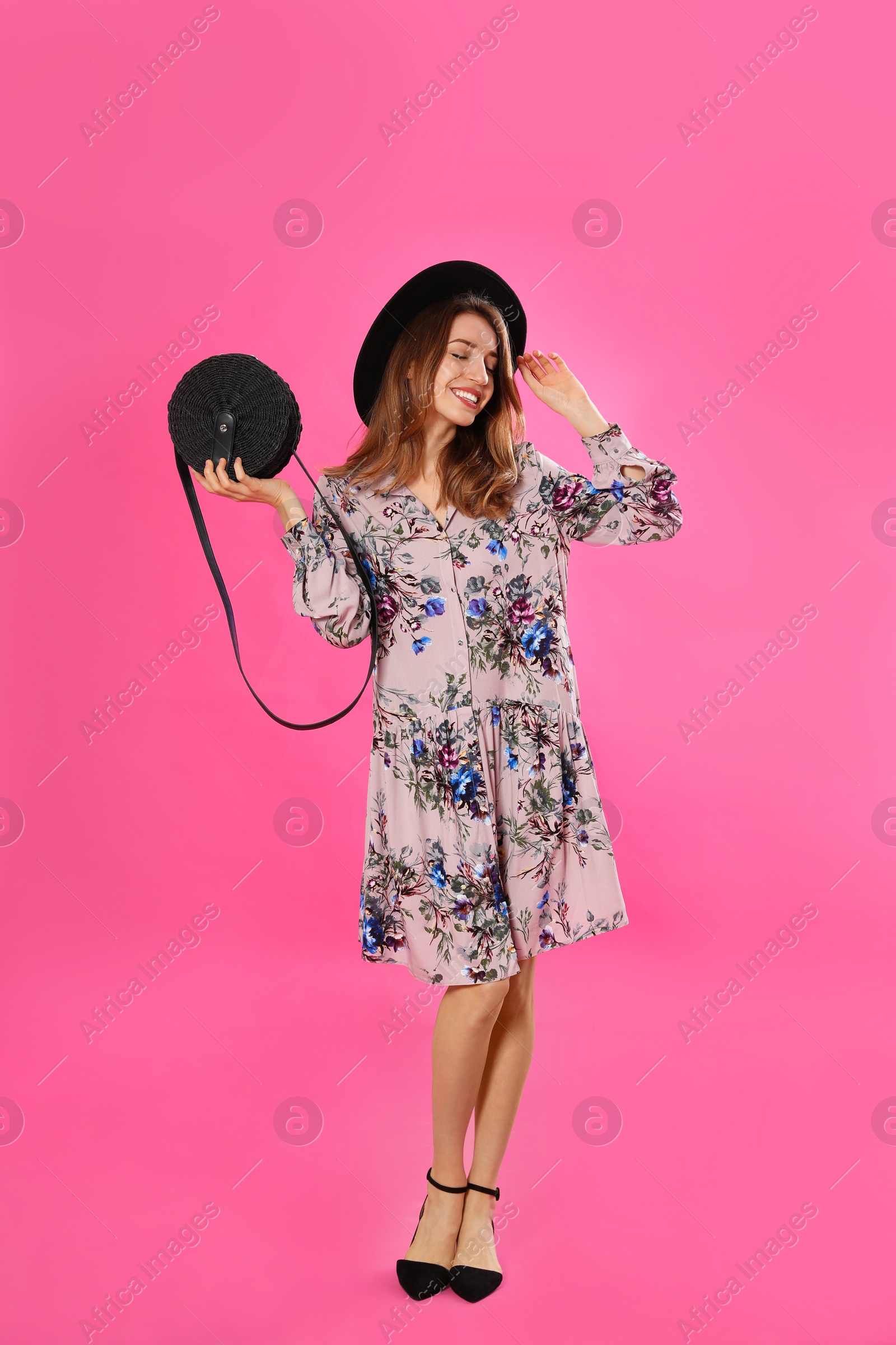 Photo of Young woman wearing floral print dress with stylish handbag and hat on pink background