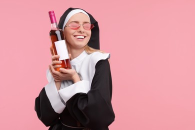 Photo of Happy woman in nun habit and sunglasses holding bottle of wine on pink background. Space for text