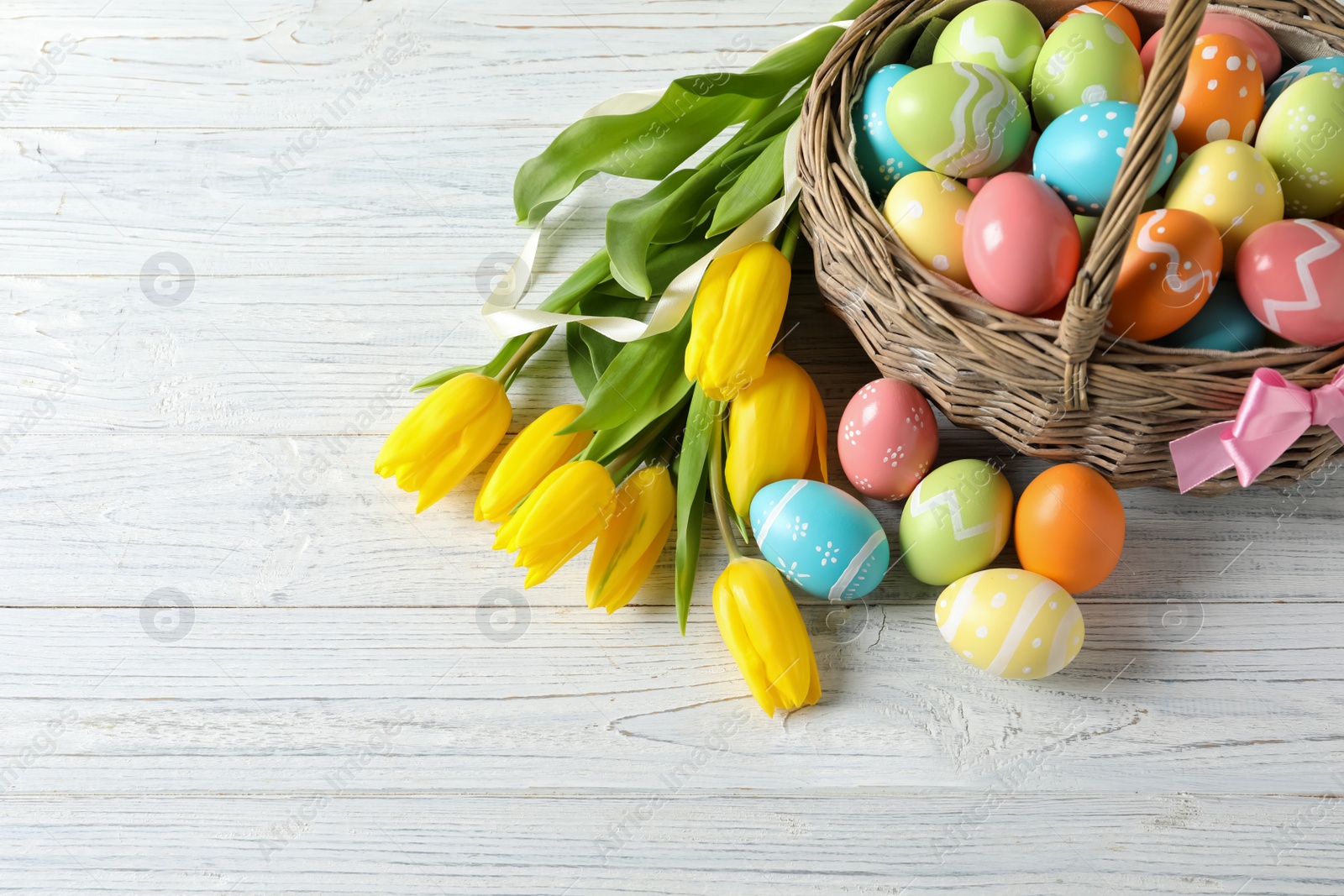 Photo of Wicker basket with painted Easter eggs and spring flowers on wooden background, above view