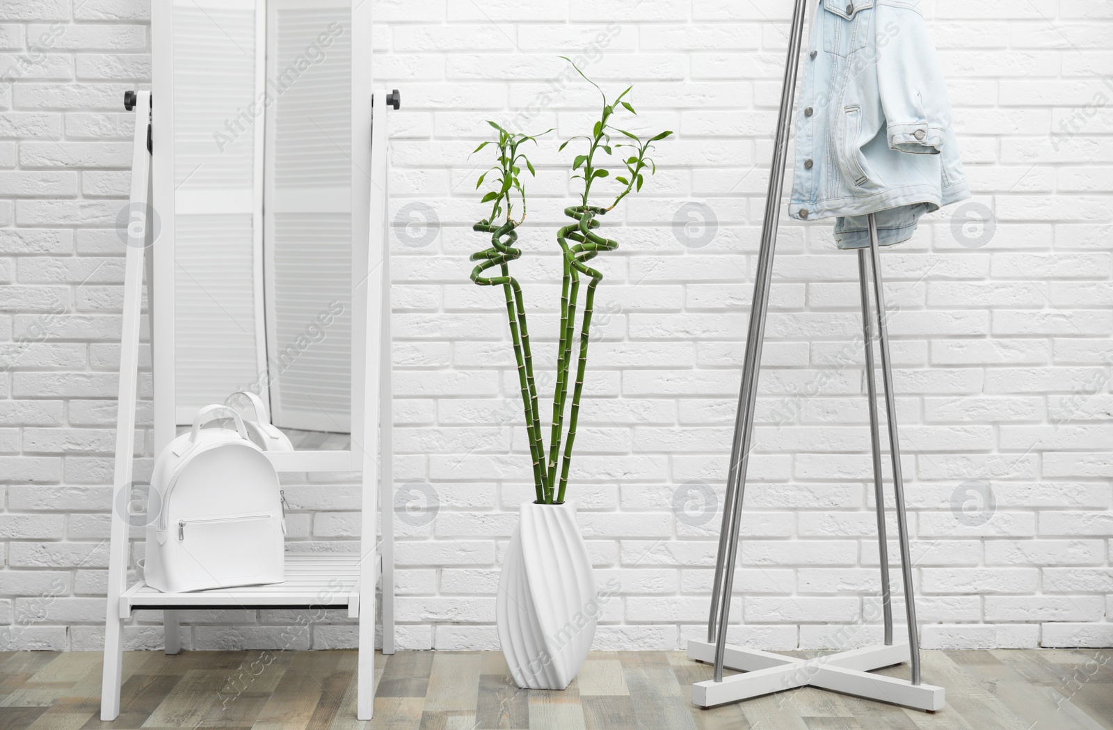 Photo of Vase with green bamboo stems, mirror and rack on floor near white brick wall in room. Interior design