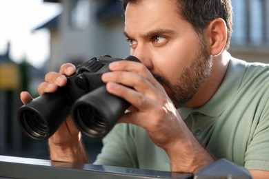 Photo of Concept of private life. Curious man with binoculars spying on neighbours over fence outdoors, closeup