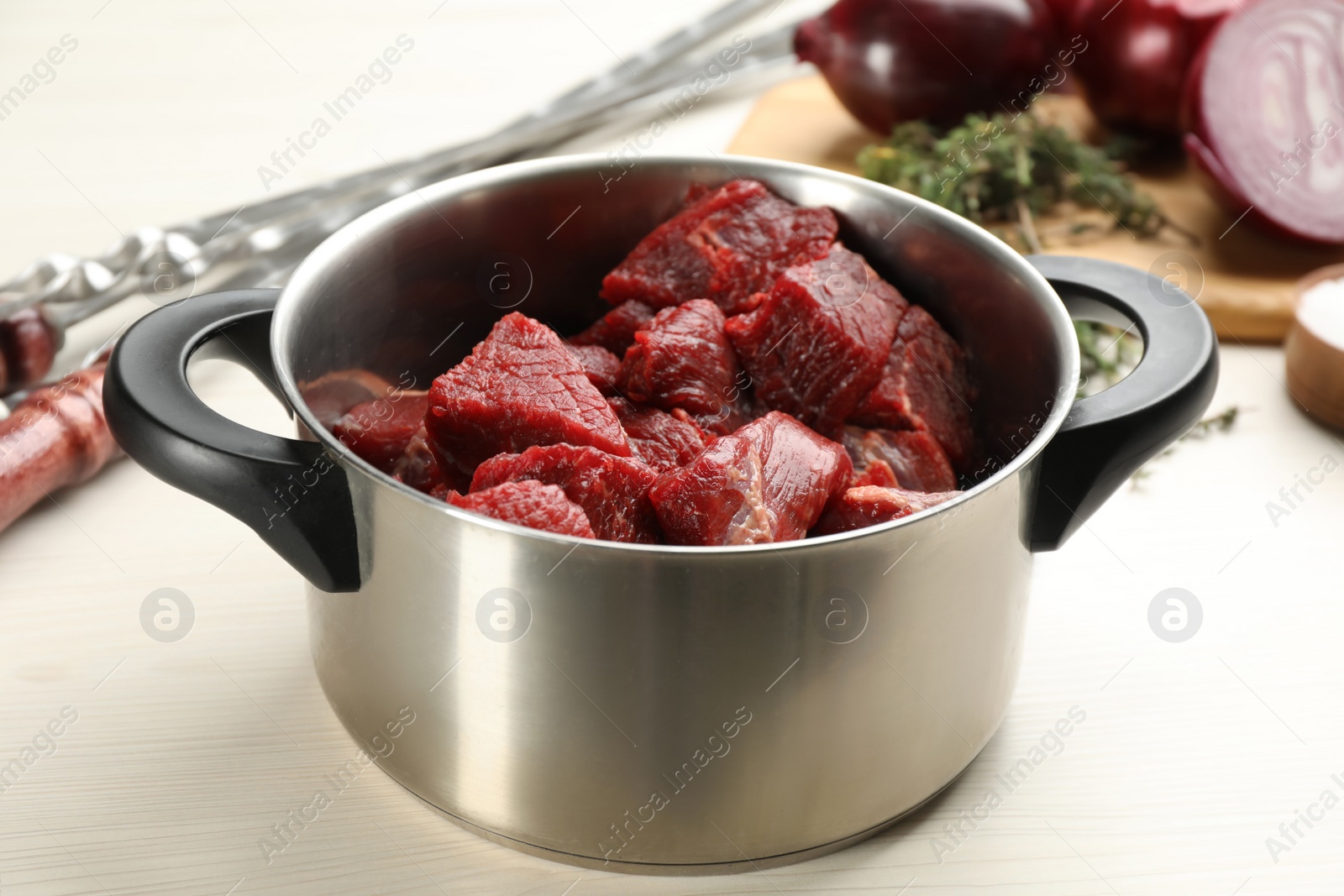 Photo of Pot with raw meat, onion and metal skewers on white wooden table