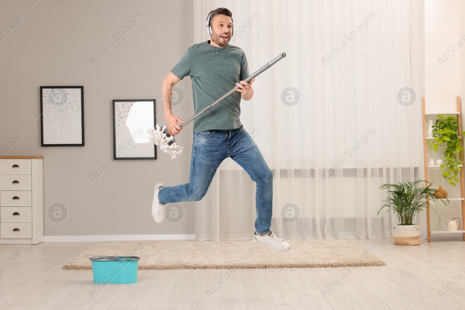 Photo of Enjoying cleaning. Man in headphones jumping with mop at home