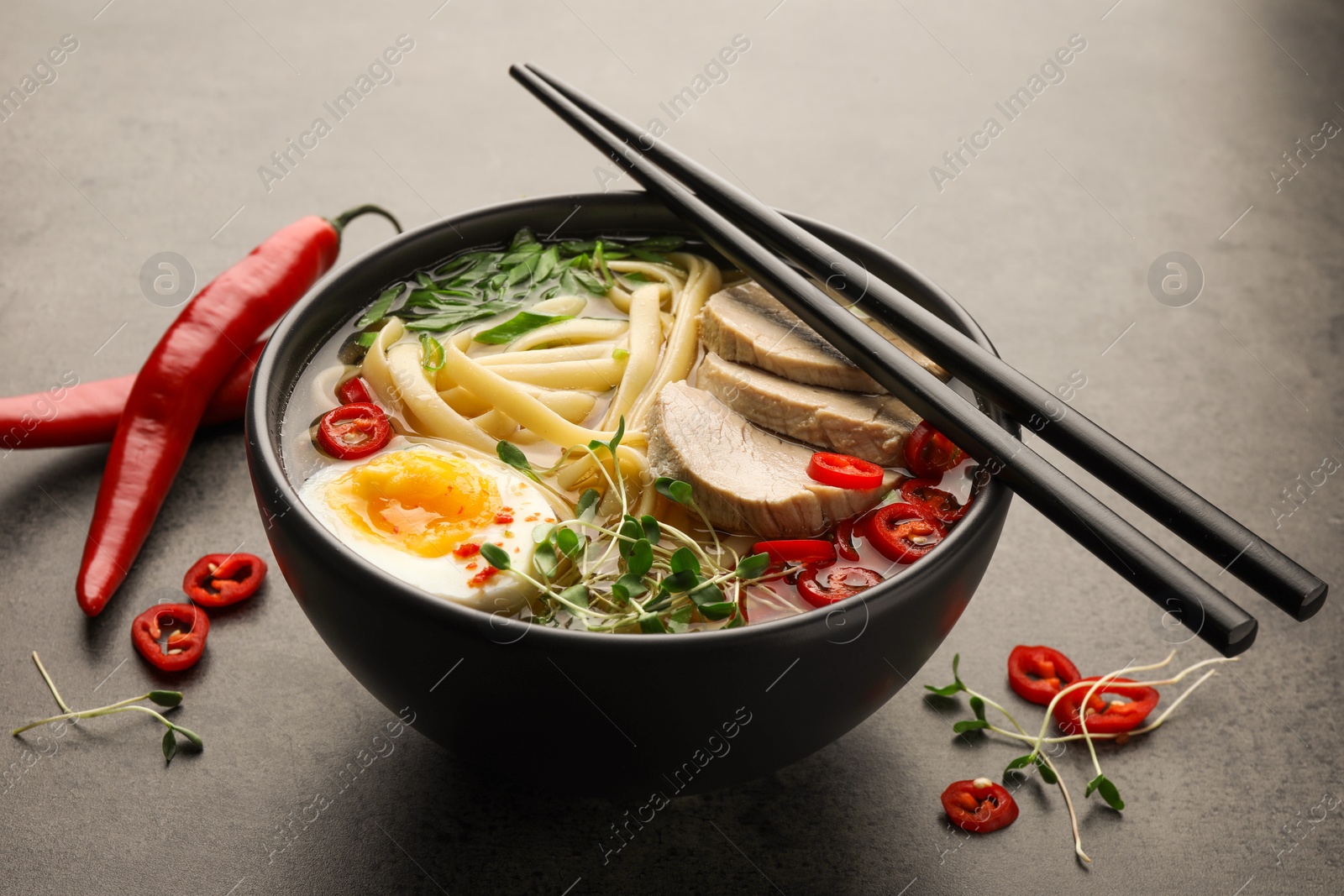 Photo of Delicious ramen with meat in bowl, chopsticks and chili peppers on grey textured table. Noodle soup