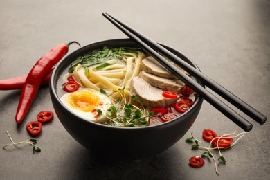 Photo of Delicious ramen with meat in bowl, chopsticks and chili peppers on grey textured table. Noodle soup