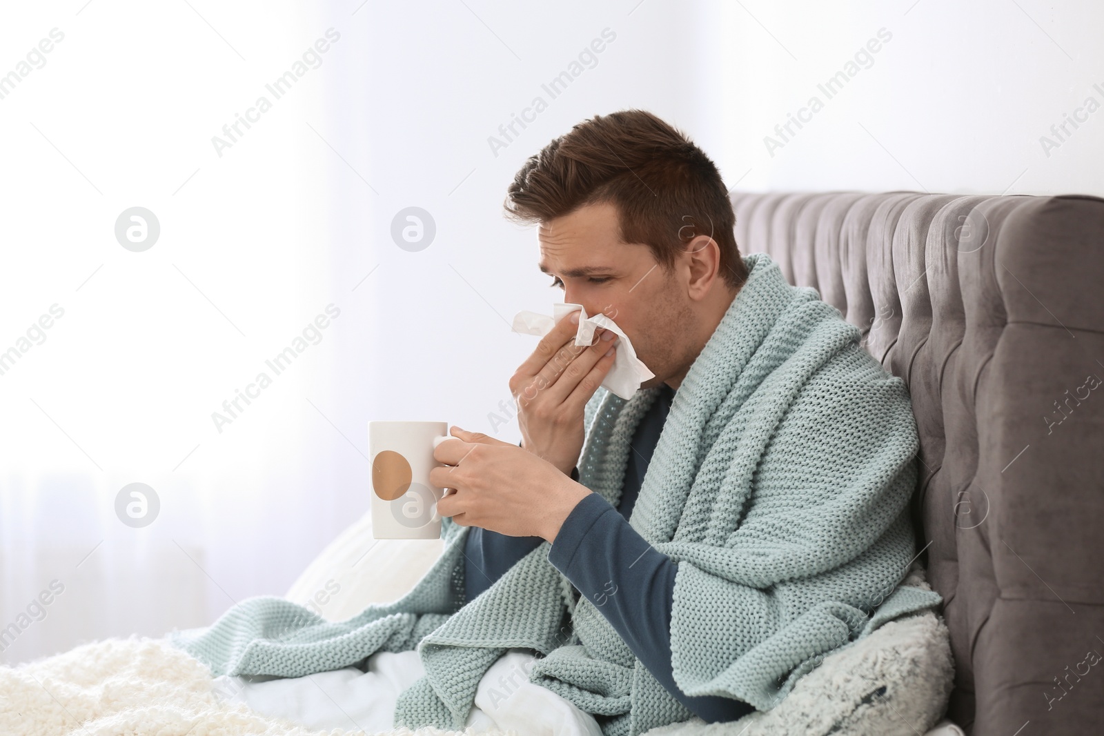 Photo of Sick young man with cup of hot drink and tissue suffering from cold in bed