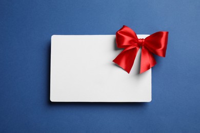 Photo of Blank gift card with red bow on blue background, top view