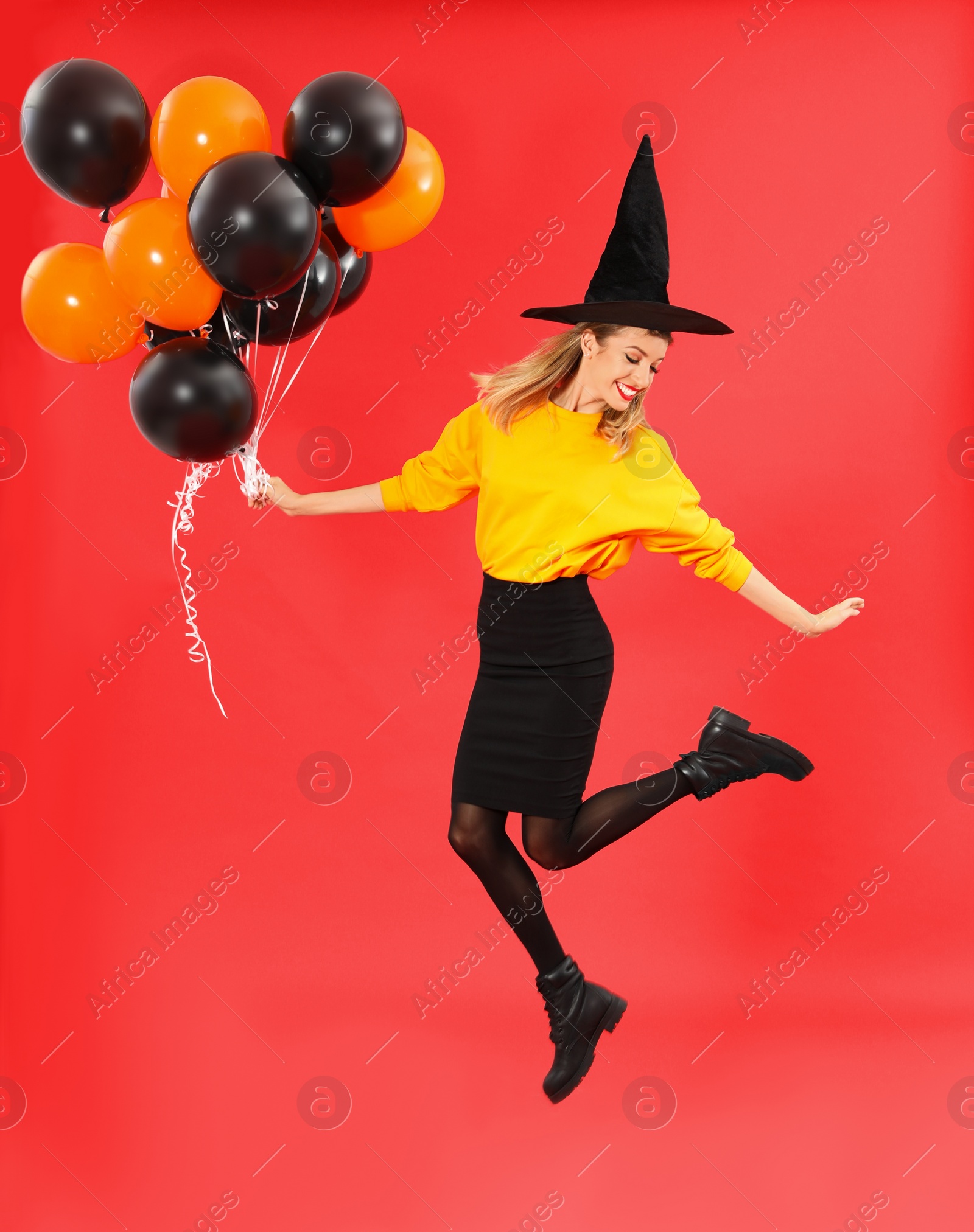 Photo of Beautiful woman wearing witch costume for Halloween party jumping with balloons on red background