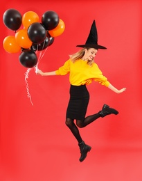 Beautiful woman wearing witch costume for Halloween party jumping with balloons on red background