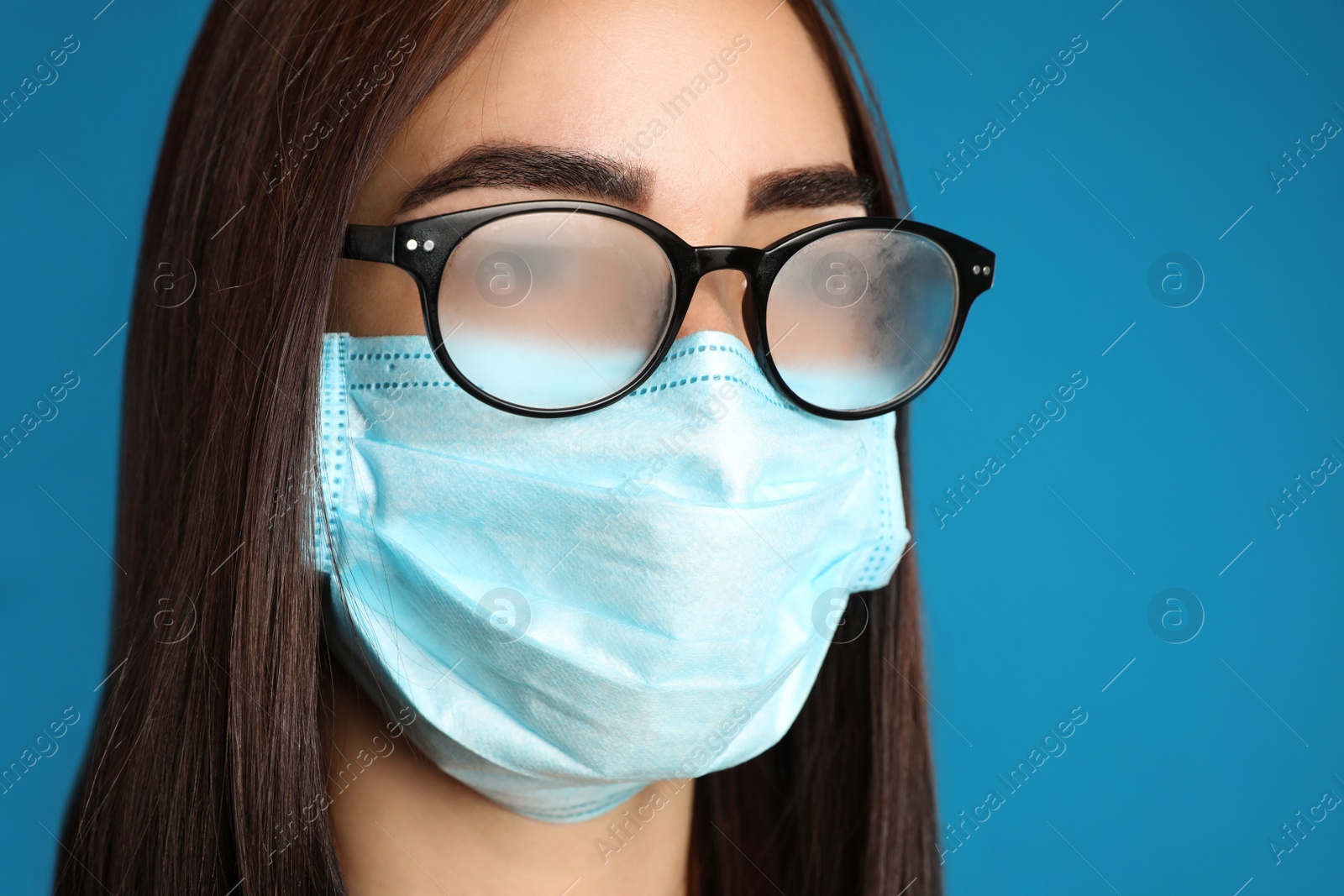 Photo of Young woman with foggy glasses caused by wearing disposable mask on blue background, closeup. Protective measure during coronavirus pandemic