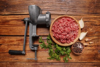 Manual meat grinder with beef mince, peppercorns, onion and parsley on wooden table, flat lay