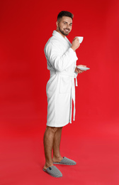 Photo of Young man in bathrobe with cup of coffee on red background