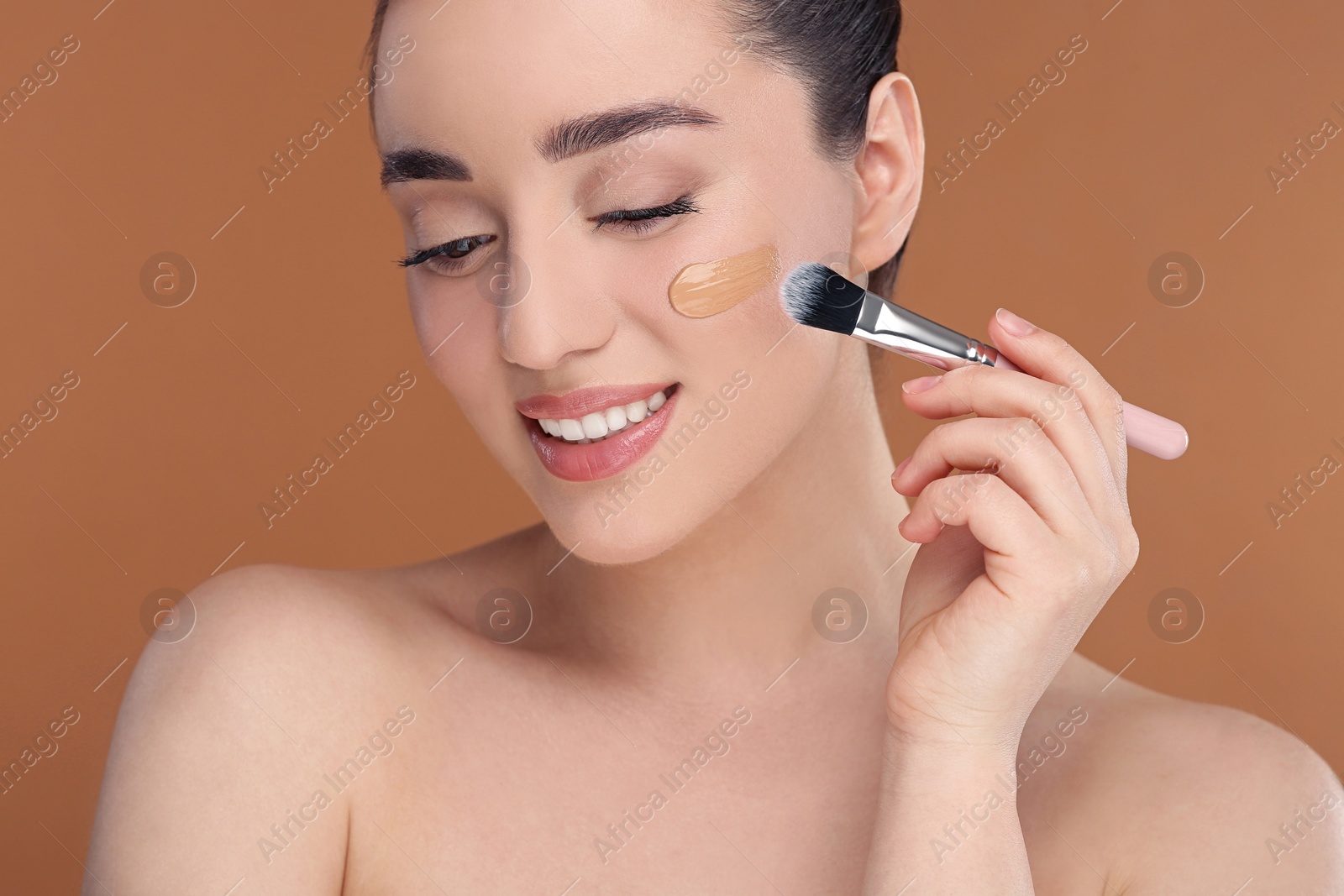 Photo of Woman applying foundation on face with brush against brown background