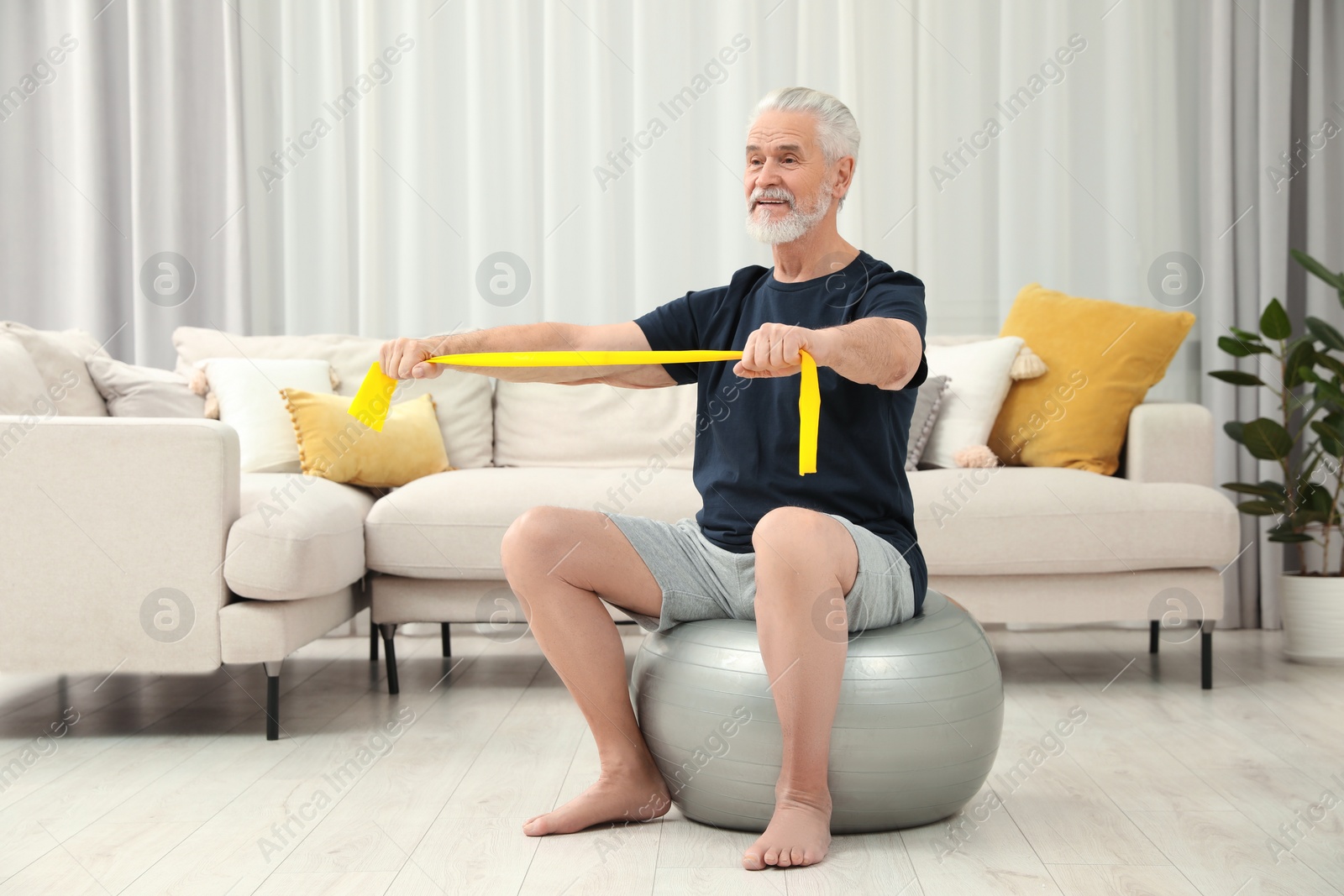 Photo of Senior man doing exercise with elastic resistance band on fitness ball at home