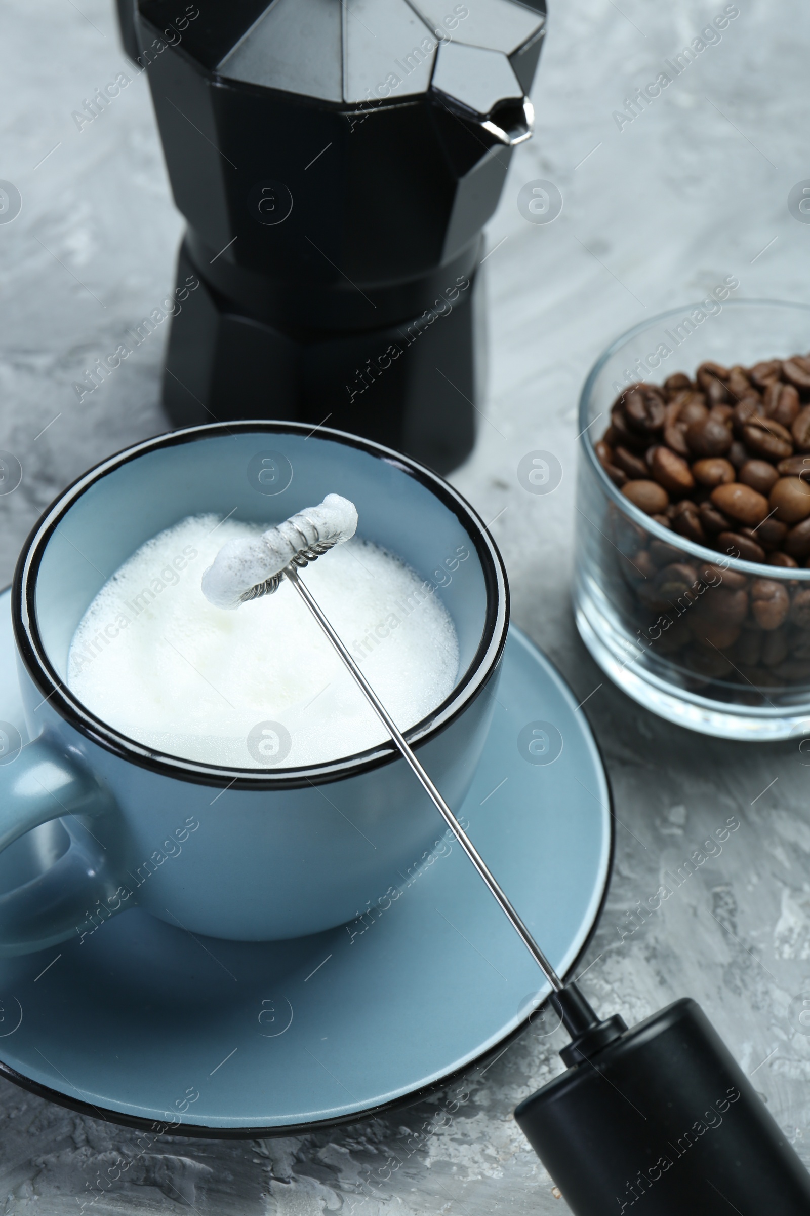 Photo of Mini mixer (milk frother), whipped milk in cup, coffee beans and moka pot on grey textured table, closeup