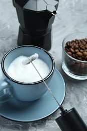 Mini mixer (milk frother), whipped milk in cup, coffee beans and moka pot on grey textured table, closeup