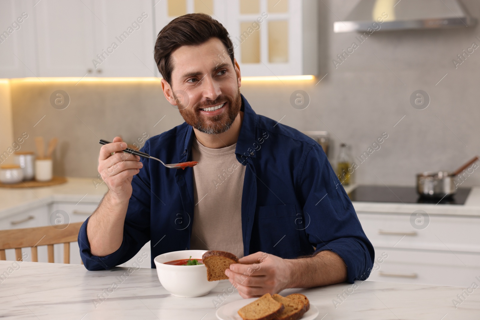 Photo of Man eating delicious tomato soup at light marble table in kitchen