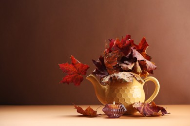 Photo of Teapot with autumn leaves and burning candle on table against brown background, space for text