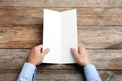 Young man holding blank brochure at wooden table, top view. Mock up for design