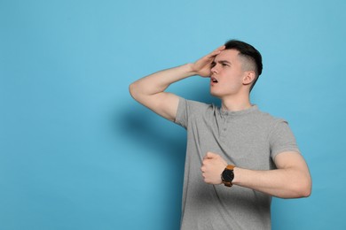 Emotional young man checking time on light blue background, space for text. Being late concept