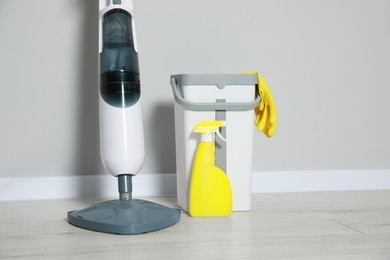 Modern steam mop, bucket with gloves and spray of cleaning product on floor near grey wall, space for text