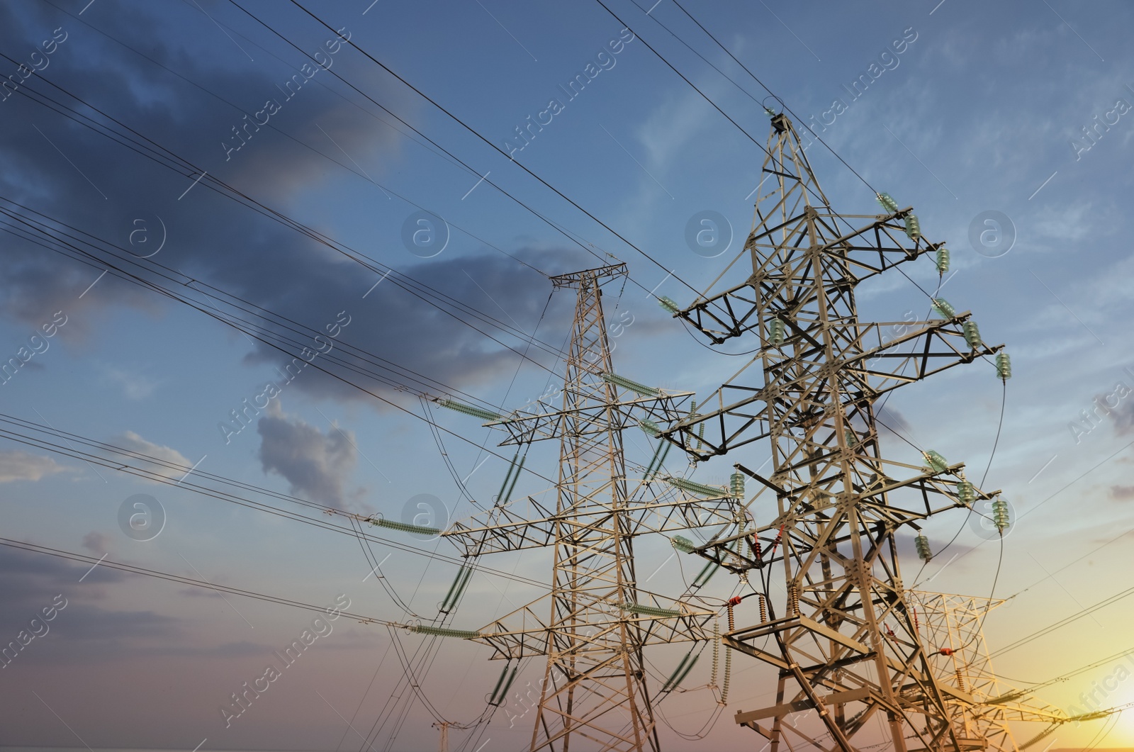 Photo of High voltage towers against cloudy sky at sunset, low angle view