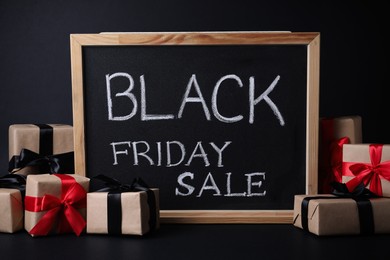 Photo of Chalkboard with words Black Friday Sale and gift boxes on dark background