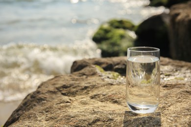Photo of Glass of fresh water on stone near sea, space for text