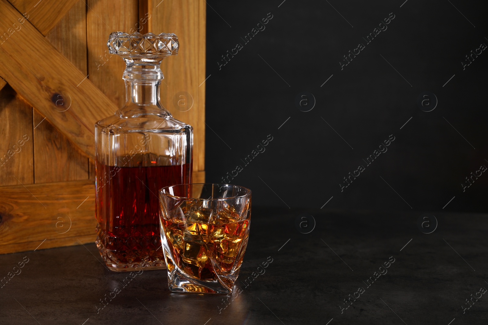 Photo of Whiskey in glass and bottle near wooden crate on dark table against black background. Space for text