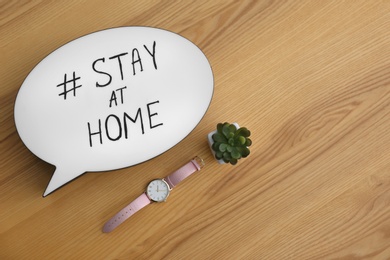 Photo of Houseplant, wristwatch and speech bubble with hashtag STAY AT HOME on wooden background, flat lay. Message to promote self-isolation during COVID‑19 pandemic