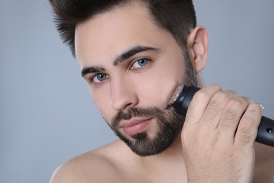 Handsome young man shaving with electric trimmer on grey background, closeup