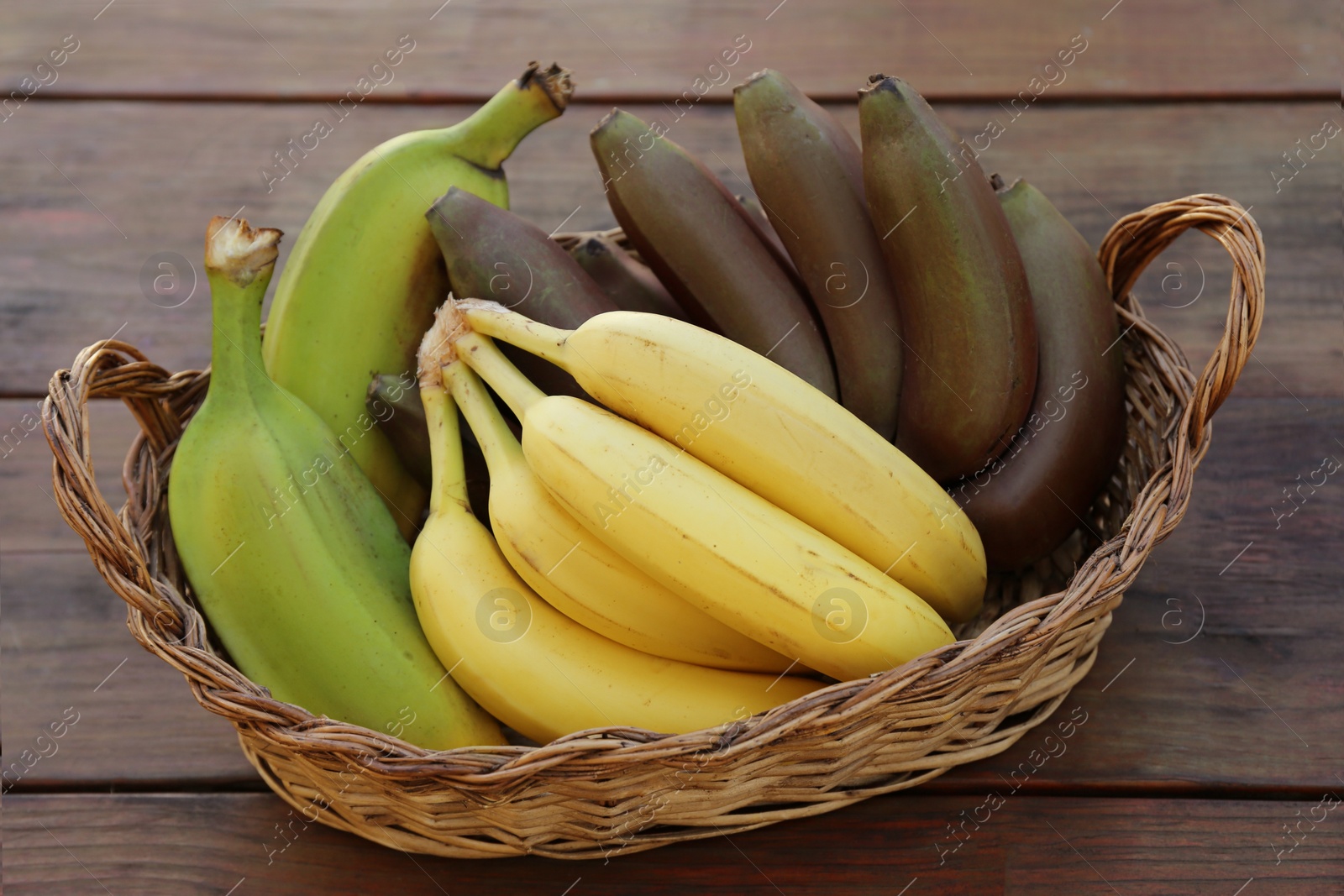 Photo of Wicker basket with different sorts of bananas on wooden table, closeup