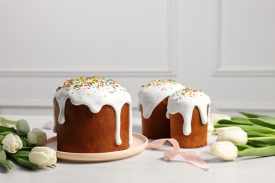 Delicious Easter cakes with sprinkles and beautiful tulips on white wooden table indoors