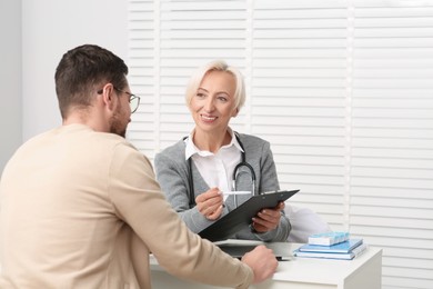 Doctor with pen and clipboard consulting patient at white table in clinic