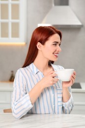 Photo of Beautiful woman with red dyed hair holding cup of drink at white table in kitchen