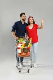 Photo of Young couple with shopping cart full of groceries on grey background