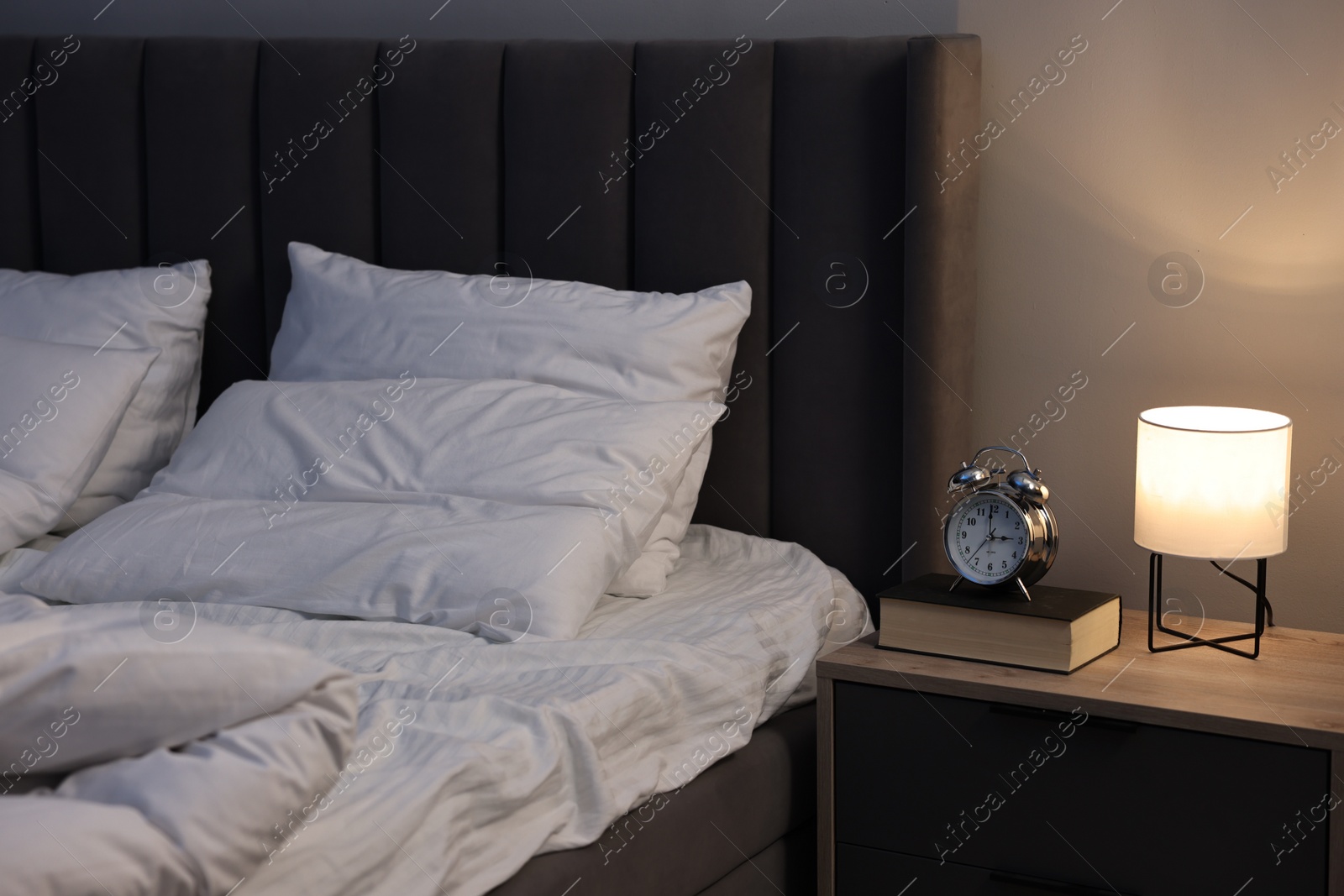 Photo of Nightlight, alarm clock and book on bedside table near bed indoors