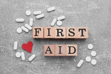 Words First Aid made of wooden cubes, pills and cardboard heart on light grey table, flat lay