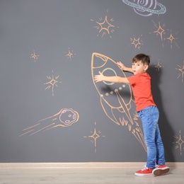 Cute little child playing with chalk rocket drawing on grey wall