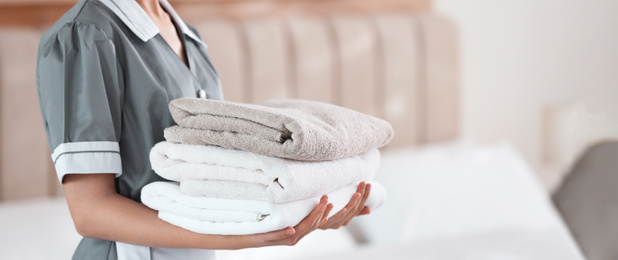 Young chambermaid holding stack of fresh towels in bedroom, closeup. Banner design