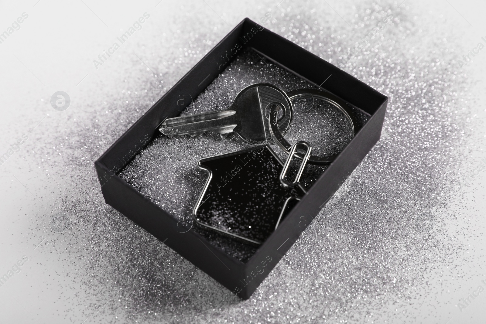 Photo of Key with trinket in shape of house, glitter and gift box on light grey background, closeup. Housewarming party