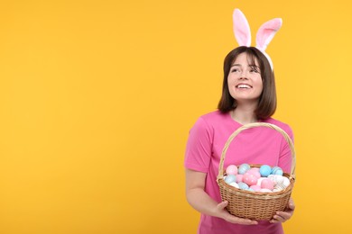 Photo of Easter celebration. Happy woman with bunny ears and wicker basket full of painted eggs on orange background, space for text