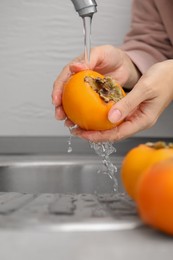 Photo of Woman washing delicious ripe juicy persimmons under tap water indoors, closeup