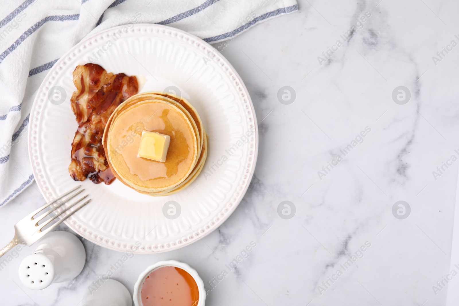 Photo of Delicious pancakes with butter, maple syrup and fried bacon served on white marble table, flat lay. Space for text